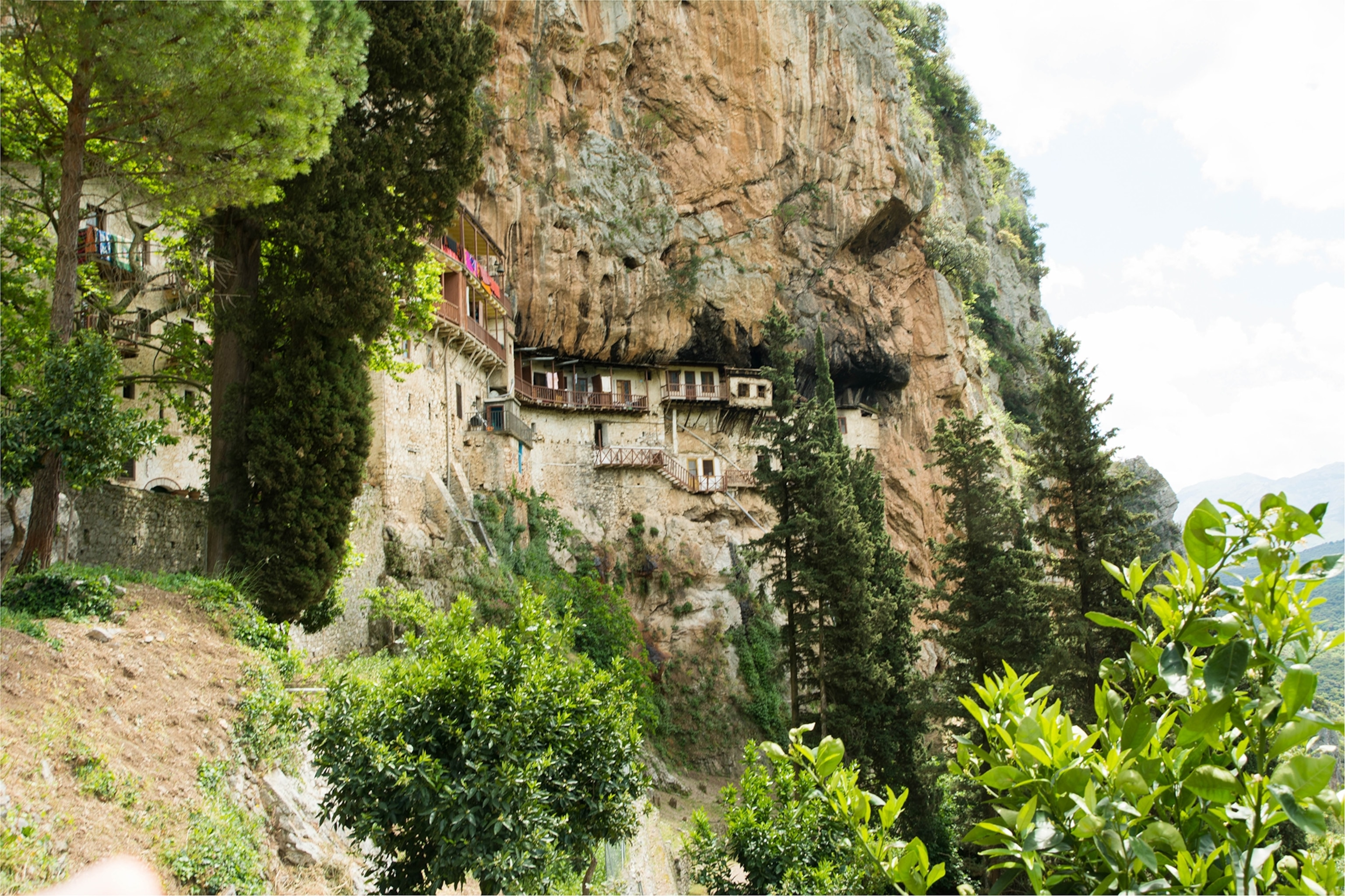 A mountain trail leading past a monastery etched into the rock, surrounded by greenery.