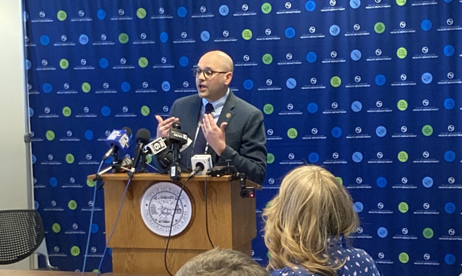 A man in a suit speaks at a podium with microphones in a room. Three people sit at a table listening, with a blue patterned background behind him.