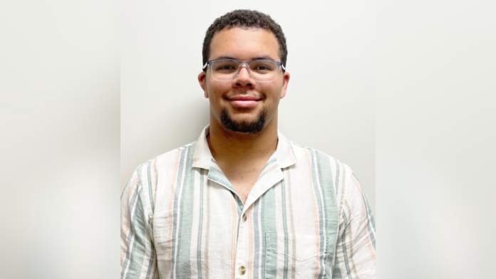 A man wearing glasses and a striped short-sleeve shirt stands against a plain white background, looking at the camera.