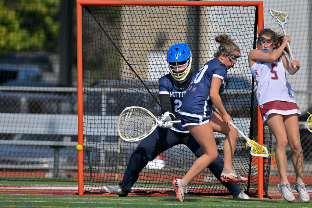 Two Mattituck Tuckers players, one field player and one goaltender, and one opponent in a maroon and white uniform, all vie for the ball.