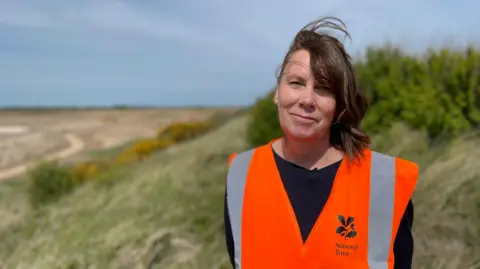 BBC A woman is standing on a grassy, sandy bank looking into the camera. Her dark, shoulder-length hair is slightly windswept and she is wearing an orange hi-vis vest.