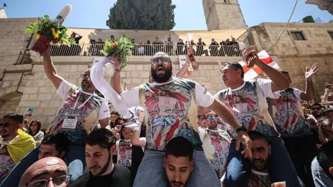 Getty Images Crowds of people outside in Jerusalem. There are four men sitting on the shoulders of other men. They are wearing matching t-shirts depicting Jesus, with Arabic inscription.