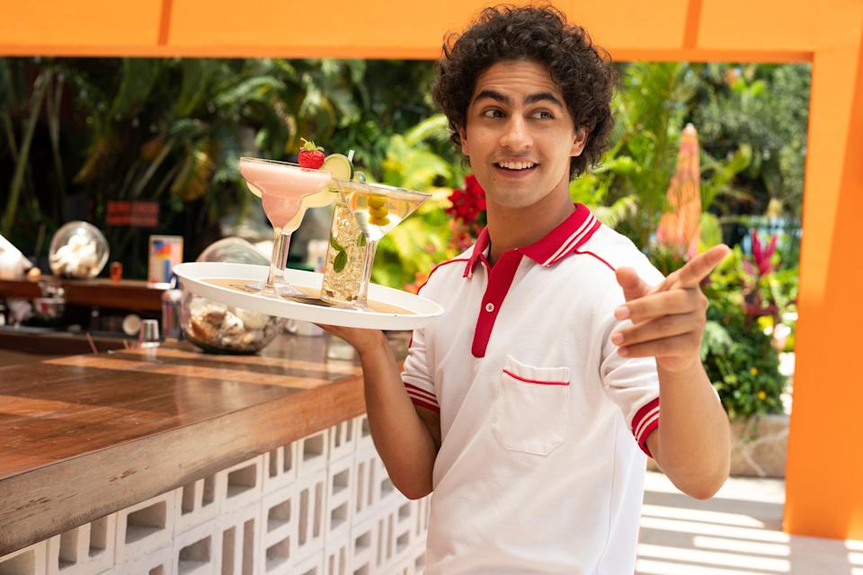 A still of "Acapulco" season one showing a man in a white and red top holding a tray of different alcoholic drinks.