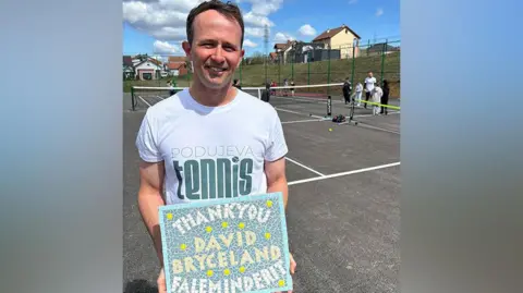 Alice Carroll David Bryceland with dark brown hair wearing a white Podujeva Tennis t-shirt holding a mosaic tile which reads 'Thank you David Bryceland Faleminderit' (translates as thank you) standing on the newly opened tennis court with children playing behind him. 