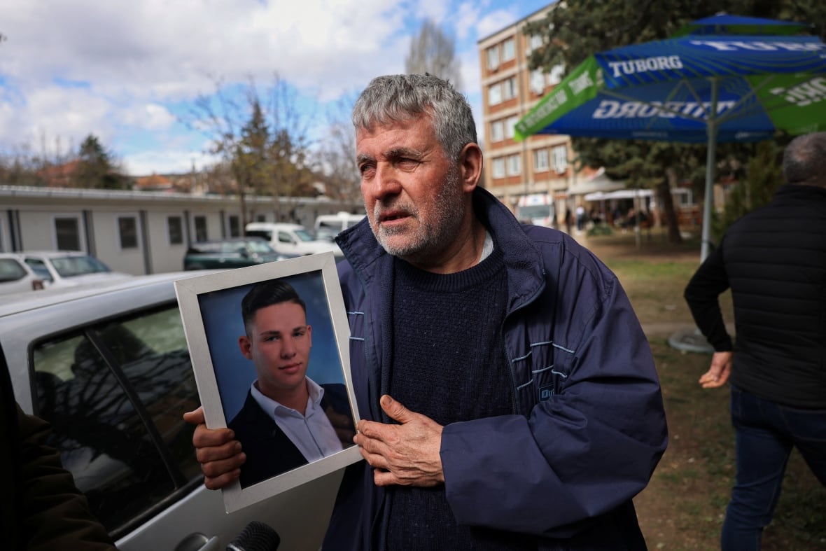 A bearded, grey-haired man in a coat outdoors holds up a framed photo of a younger man.