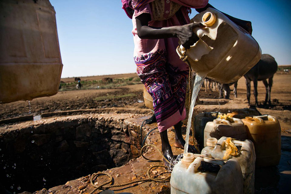 In teh left of the image a circular opening to a water well can be seen, the land beyond is arid brown and green under a blue sky. The lower two thirds of a woman can be seen in the right of the image, she is pouring water into jerry cans.
