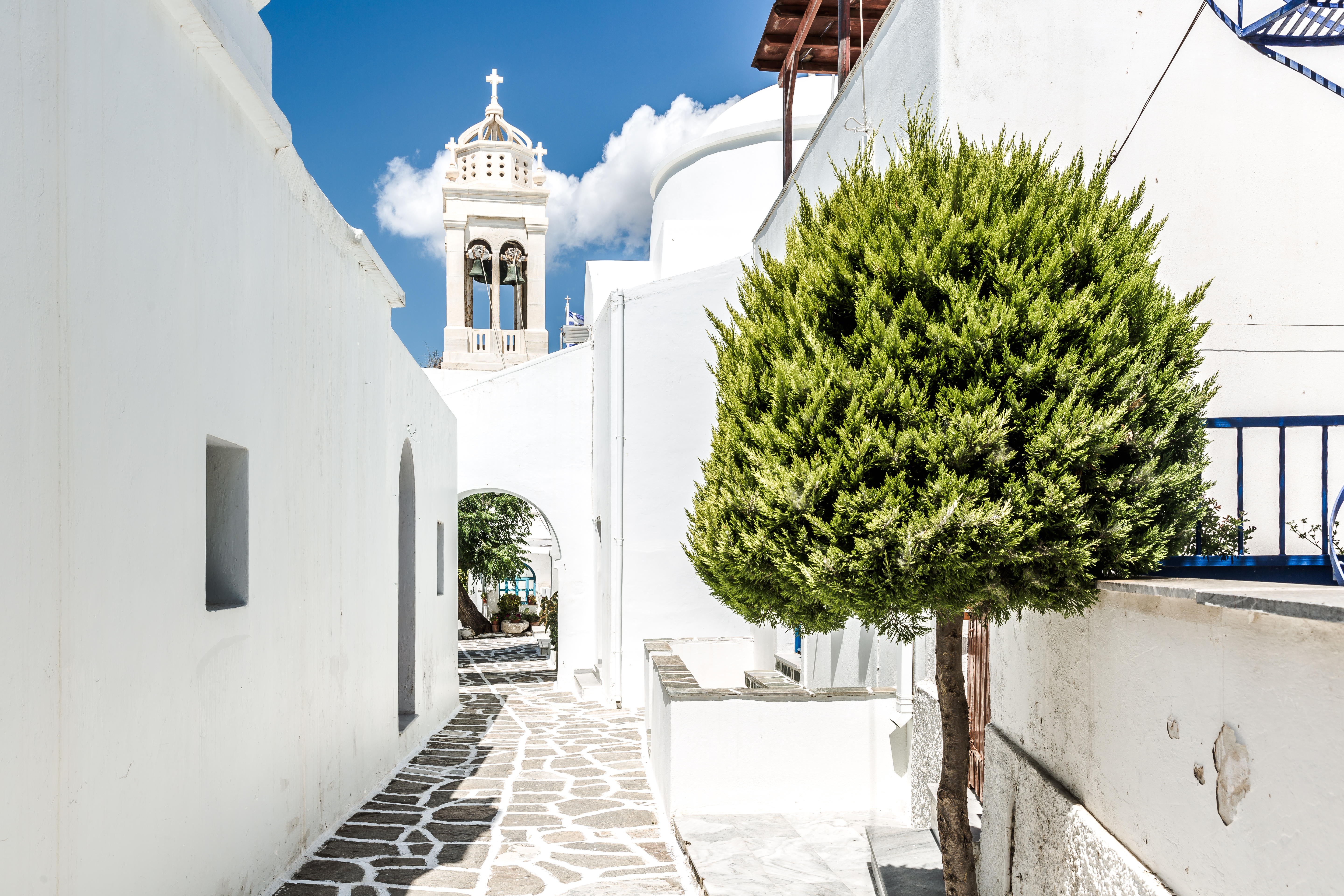 Whitewashed alleyway in Prodomos village, Paros, Greece, with a church bell tower visible.