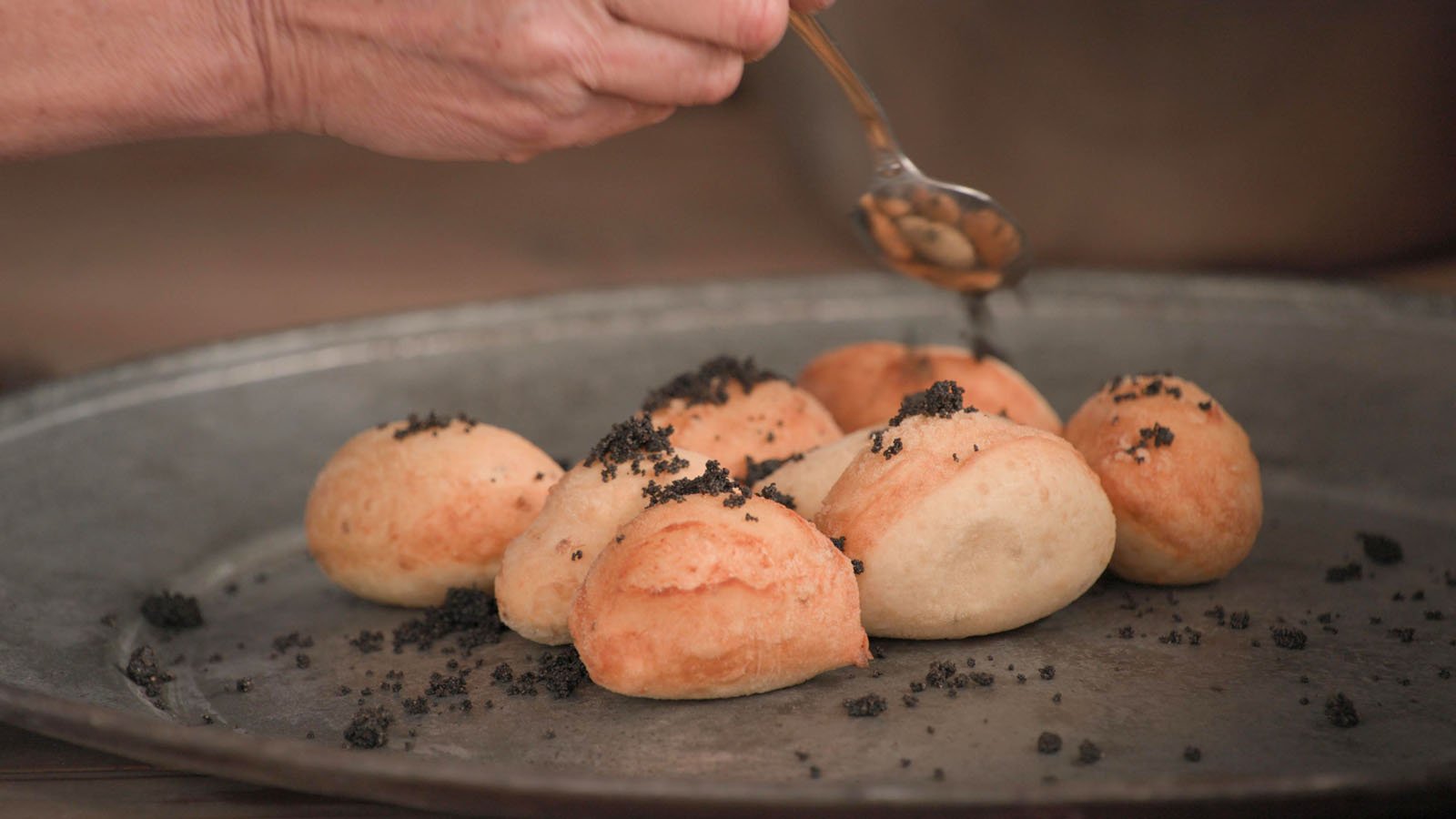 A hand uses a spoon to sprinkle a dark topping over a tray of freshly baked, golden-brown bread rolls.