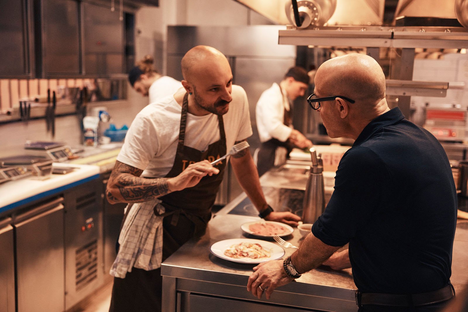 A chef in an apron talks with a man in glasses at a kitchen counter with plated food, while other staff work in the background of a modern, professional kitchen.