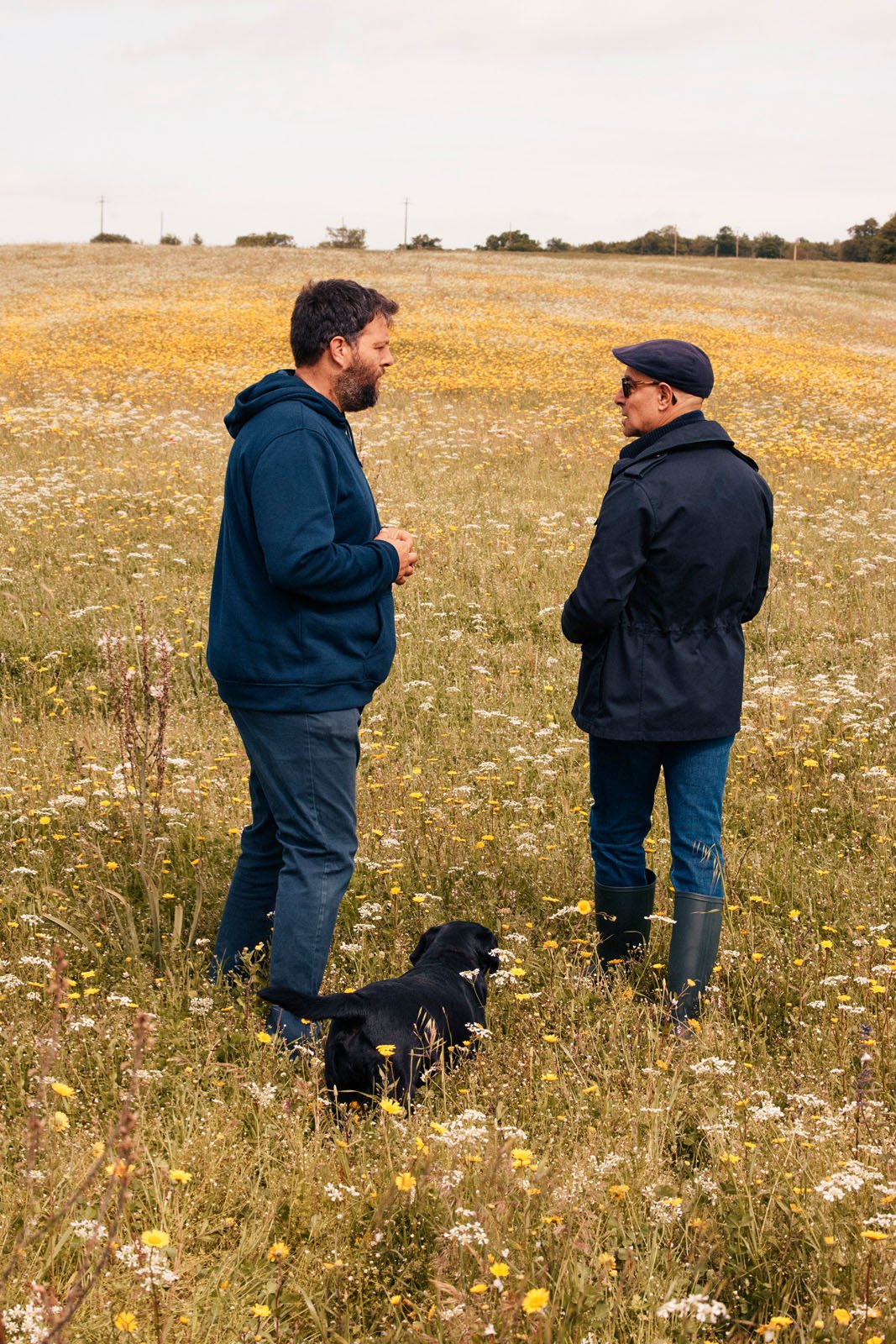 Two men stand and talk in a field of wildflowers, both dressed in jackets and jeans. A black dog sits at their feet. The sky is mostly cloudy, and the landscape extends into the distance.