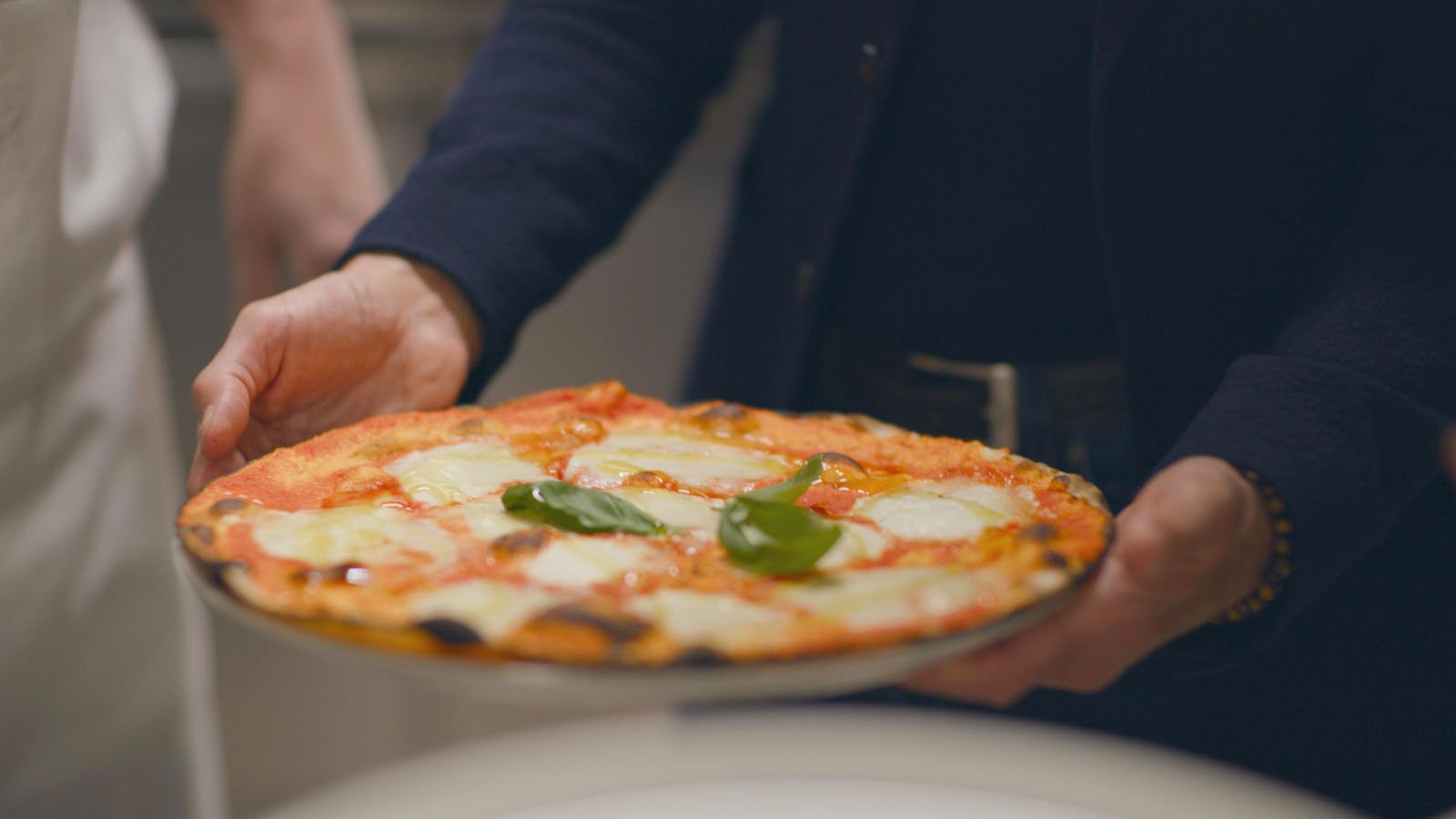 A person holds a fresh pizza topped with melted cheese, tomato sauce, and basil leaves. The pizza is being presented on a plate, with hands visible and background slightly out of focus.