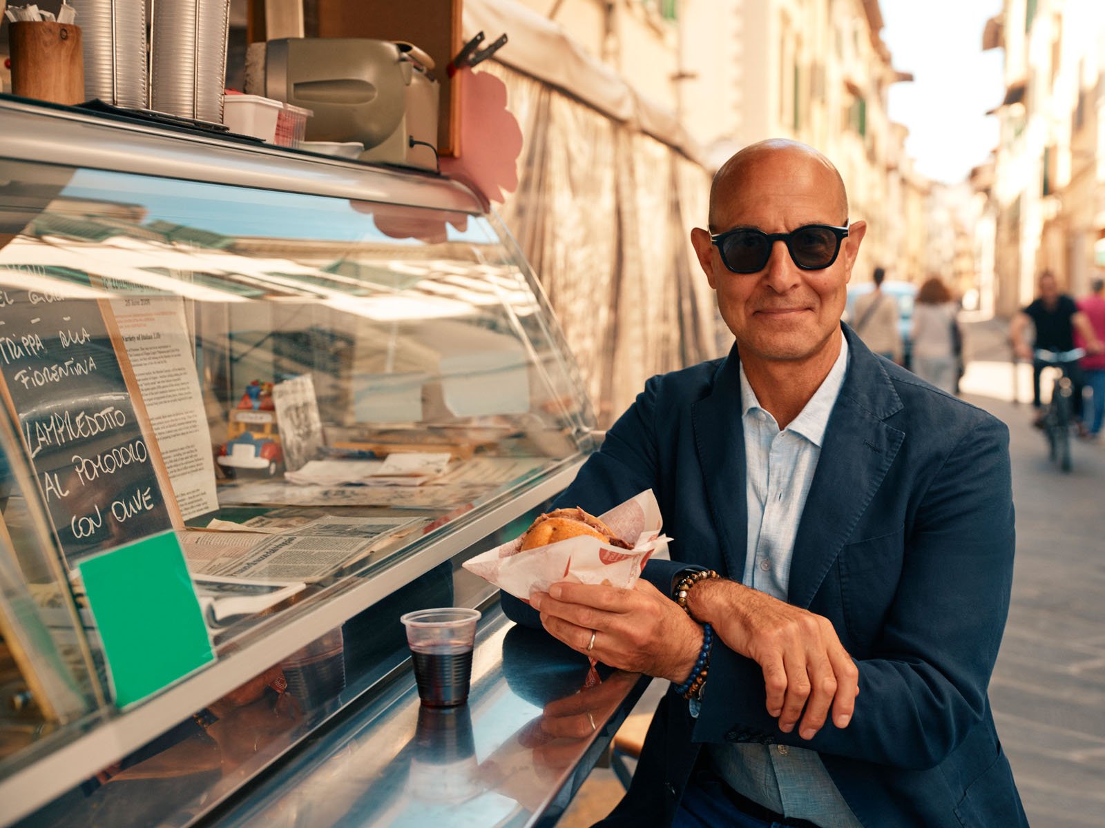 A bald man in sunglasses sits at an outdoor food stand, smiling and holding a sandwich with a drink beside him. The scene takes place on a sunny street with people walking and biking in the background.