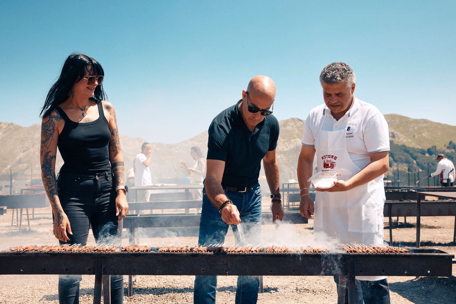 Three people stand outdoors grilling skewers over smoky barbecue grills with mountains in the background; one man seasons the food while another in a white apron and a woman watch and smile.