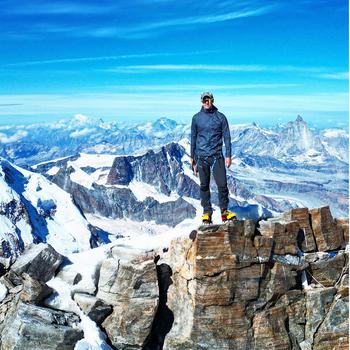 Russell stands atop a mountain with snowy peaks and blue sky in the background.