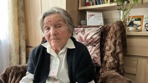 98-year-old Wanda Traczyk-Stawska looks directly into the camera. She is sitting on an armchair wearing an embroidered blouse and a cardigan. 