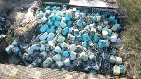 Environment Agency An aerial view of bales of rubbish which have been dumped on a farm in Lincolnshire. The blue-coloured bundles are piled up and surrounded by two agricultural buildings.