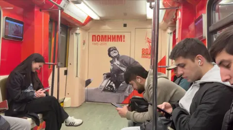 The inside of a Moscow metro carriage. The passengers are look at their phones but the back wall of the carriage has a black and white photo of a World War Two pilot and a logo celebrating the 80th anniversary of Russian victory. 