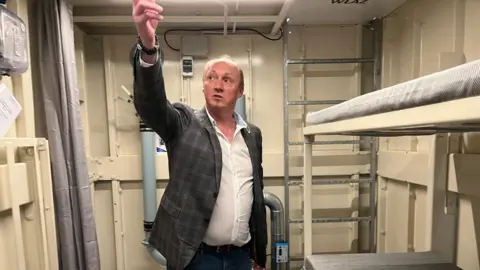 Janusz is wearing a checked jacket and jeans inside one of his bomb shelters. He is pointing to the ceiling. The shelter contains a bunk bed and a toilet with a curtain. It is clean but sparse. 