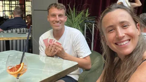 Rudi Voller Rudi and Lizzie sitting at a table in a hotel lobby. Both are smiling. Rudi is holding a deck of cards in his hands.