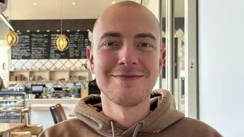 Handout A man with a very short buzz cut hairstyle. He has a stubbly beard with a nose ring and is wearing a brown hoodie. He appears to be in a restaurant. 