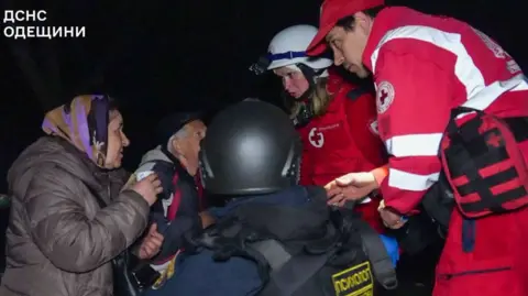 DSNS Rescuers wearing red and white high vis uniforms and helmets speak to local residents as they evacuate people following the strike.