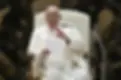 A man in white robes sits on a chair, holding papers and gesturing with his right hand. He is speaking in a formal setting with a microphone.