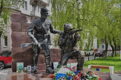 Two statues - one is a man wearing a military uniform from World War Two. He has his hand on the shoulder of a man wearing a modern military uniform, camos with a helmet, who is crouching and aiming a gun. 