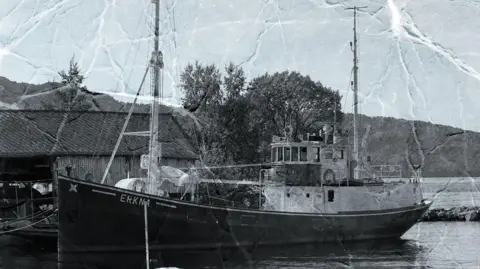 The Liberation Convoy A black and white picture of the the M/K Erkna - a medium sized boat stationed by the shore with a building nearby. The picture appears to have been scanned so you can see lots of lines where it was folded. 