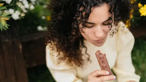 Getty Images Stock photo of a close up shot of a person with black hair eating what looks like a chocolate covered protein bar outdoors