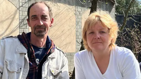 BBC A middle-aged man and woman, sitting in front of a wall and a tree, look at the camera 