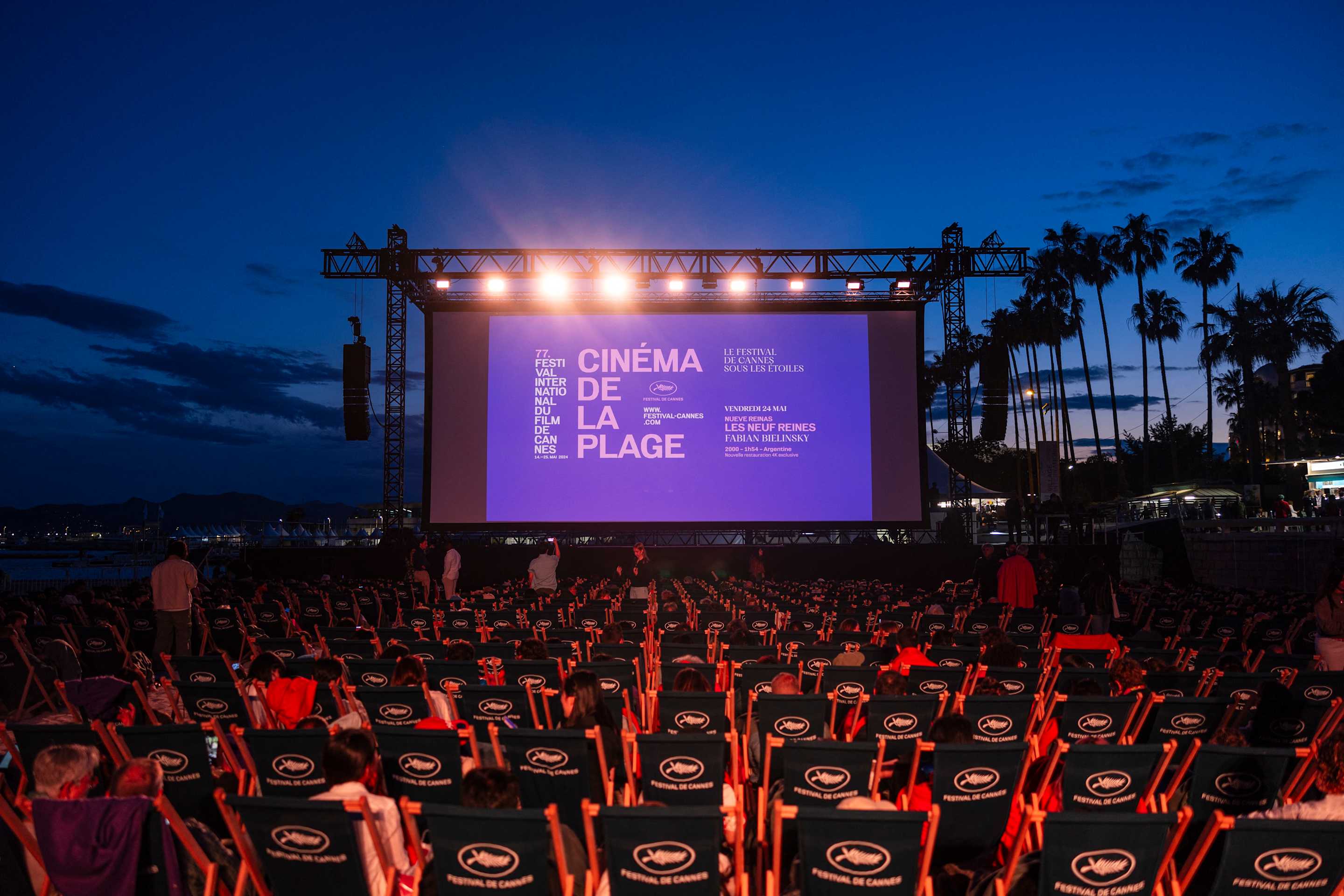 Spectators watch an outdoor movie near palm trees.