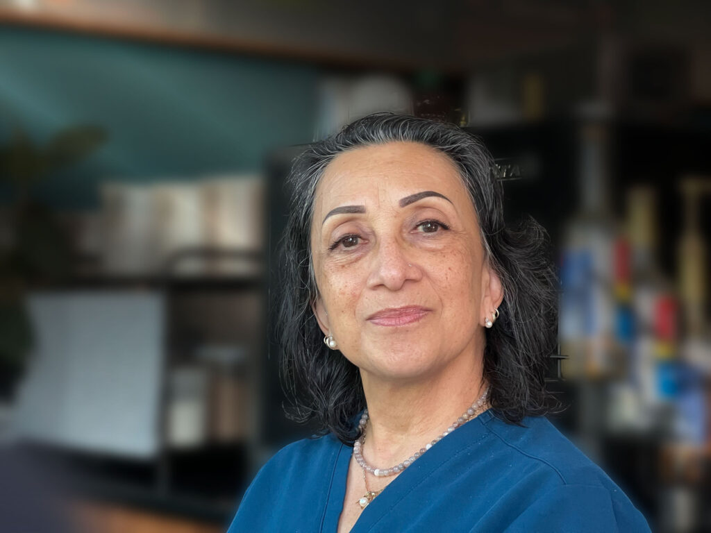 A middle-aged woman with gray hair and wearing a blue top looks at the camera with a neutral expression. The background is an indoor setting, slightly blurred.