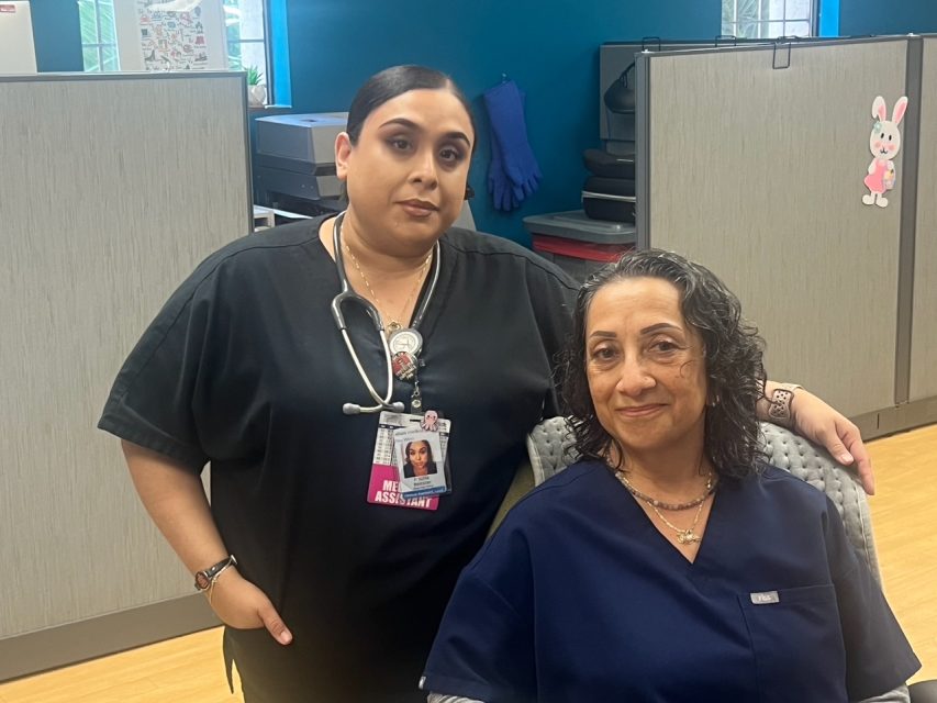Two women in medical scrubs pose in an office setting; one is standing with a stethoscope and badge, the other is seated and smiling.