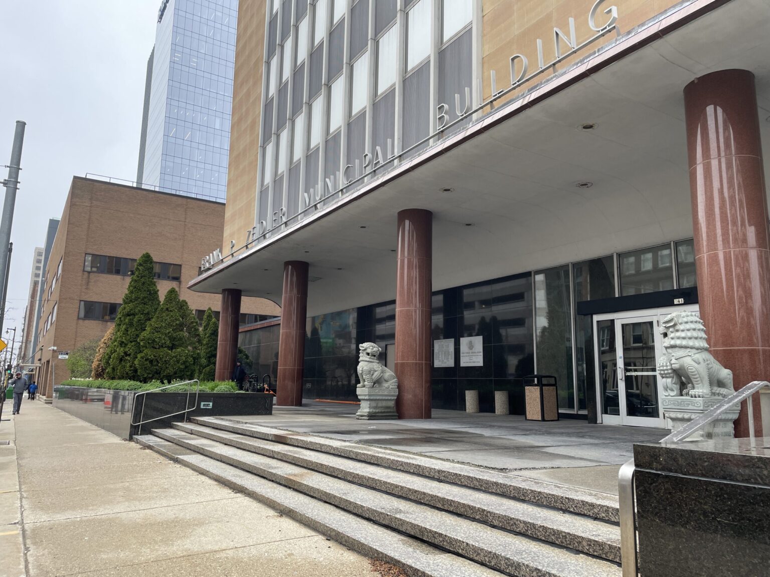 A wide sidewalk runs past the entrance of a municipal building with large columns, steps, and two stone lion statues flanking the main doors.