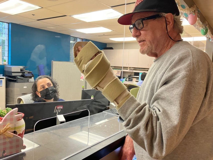An older man with a foam cast on his hand speaks to a masked receptionist at a front desk, separated by a clear barrier.