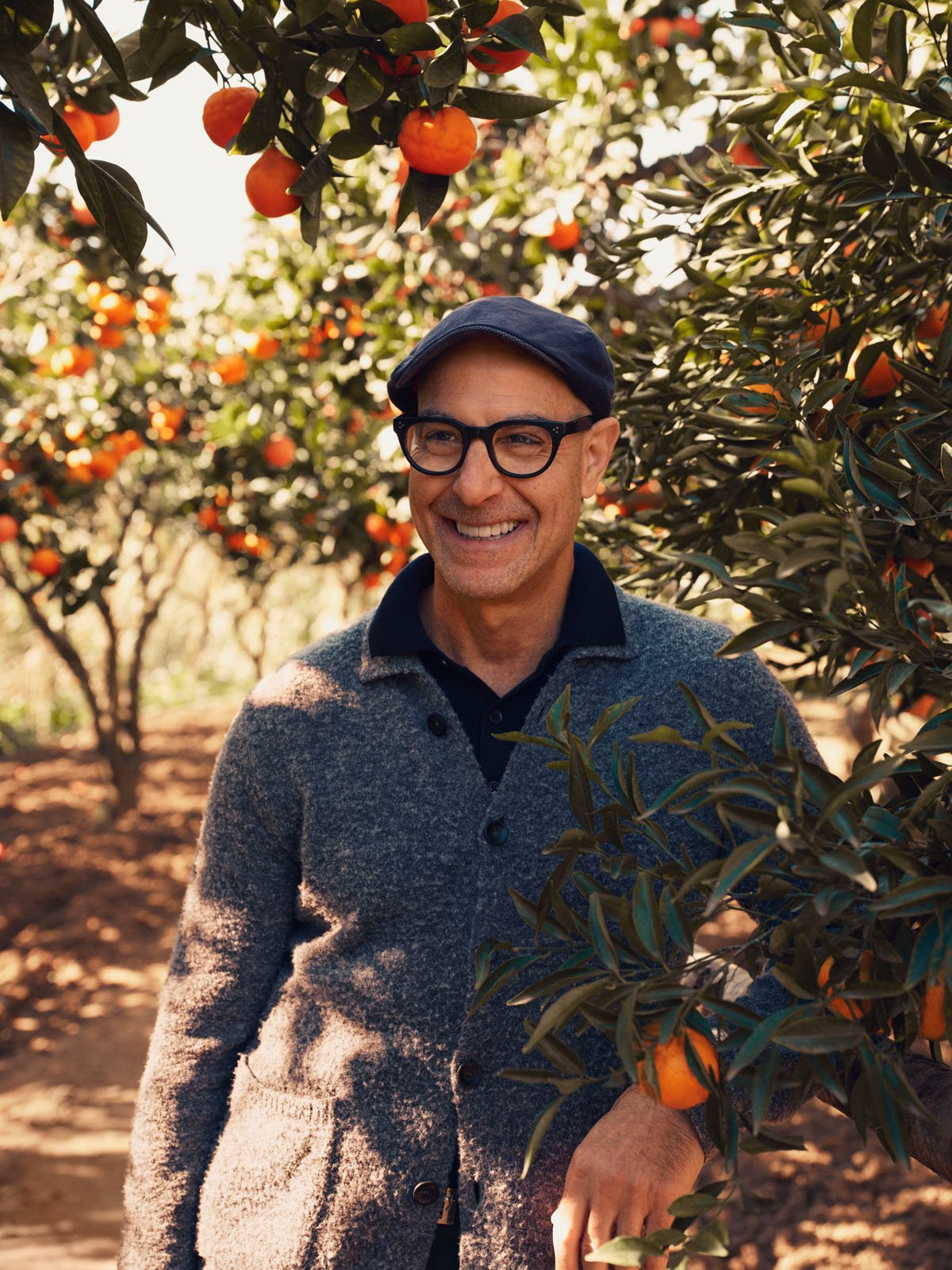 A smiling man wearing glasses, a blue flat cap, and a gray cardigan stands among orange trees laden with ripe fruit on a sunny day.