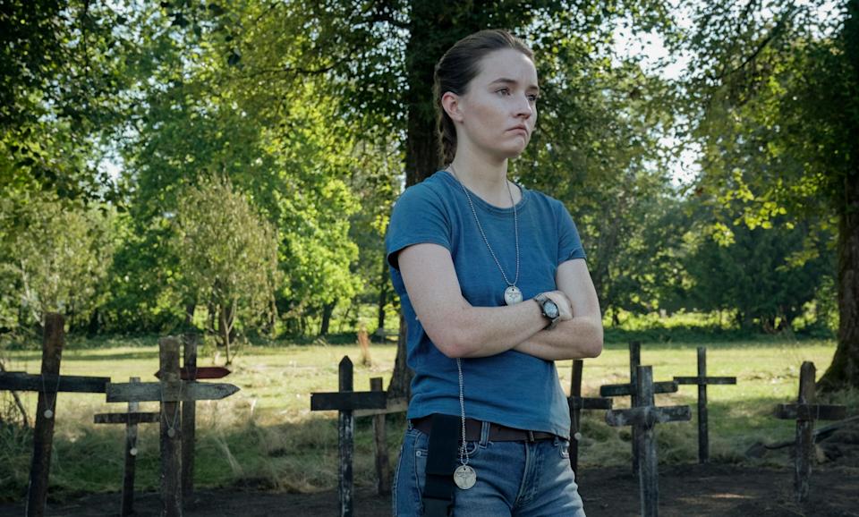 A young woman with brown hair tied back in a ponytail. She's wearing a plain blue t-shirt, blue jeans and a brown belt. There is a green and black watch on her left wrist. There is a silver medallion hanging around her neck. There are wooden crossed on the ground directly behind her, with a field that has several trees in it further in the background.