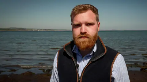 BBC A bearded man wearing a cardigan and striped shirt standing in front of the sea