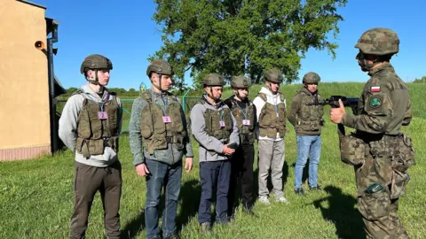 A line of six people wearing camo helmets and vests watching a soldier in full camo with a Polish flag badge show them a gun. They are standing in a field. 