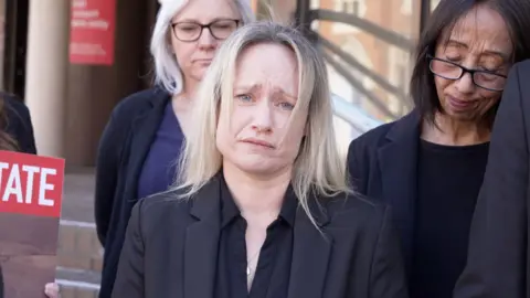 Shaun Whitmore/BBC A woman with blonde hair wearing all black. She looks devastated and is holding back tears. There are people standing behind her also looking very upset.