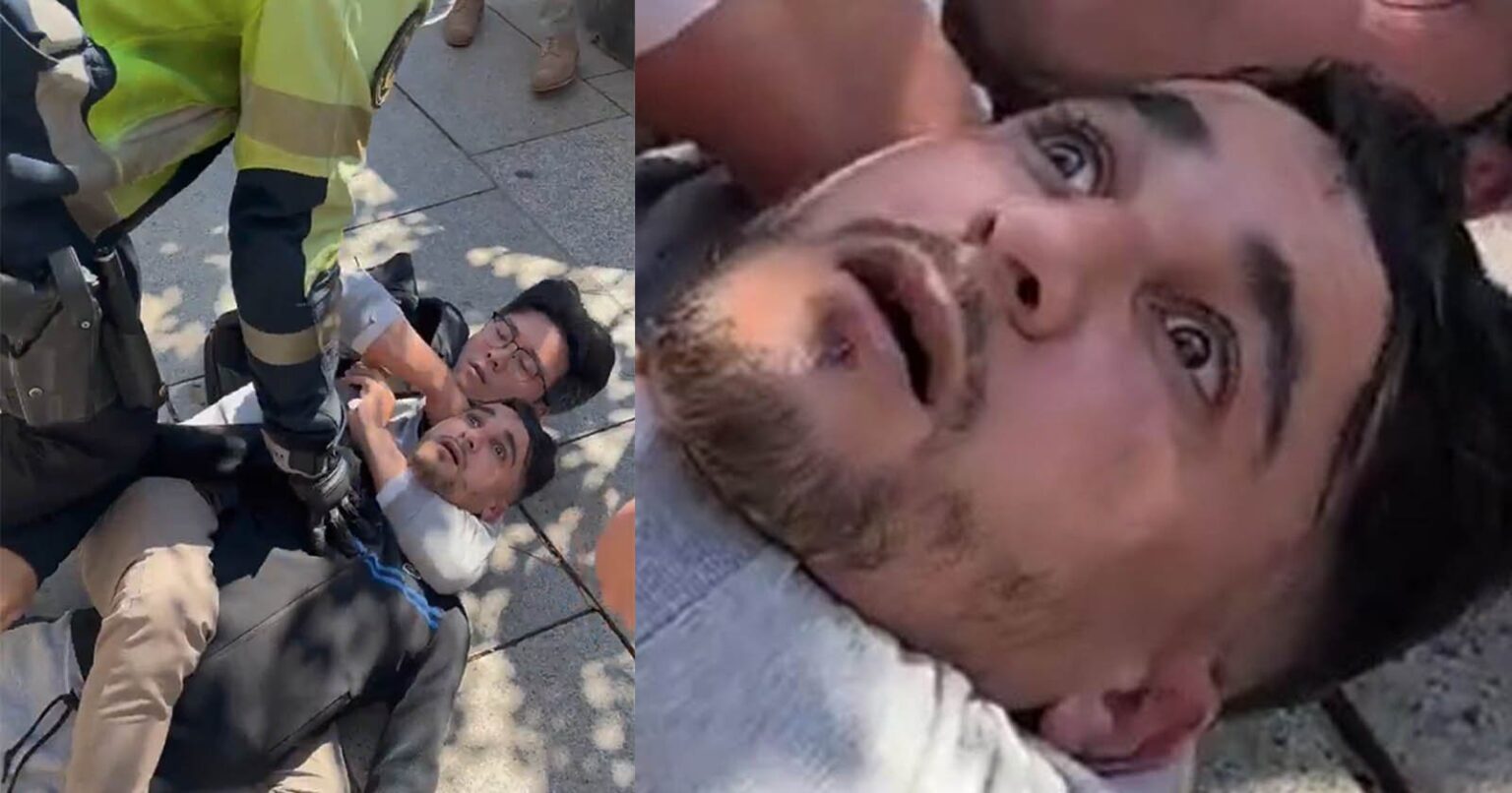 Chinese Photographer Uses Chokehold to Capture Camera Thief in Spain A close-up of a distressed man lying on the ground, being restrained by a uniformed officer, with another man beside him. The man's facial expression shows fear and shock.