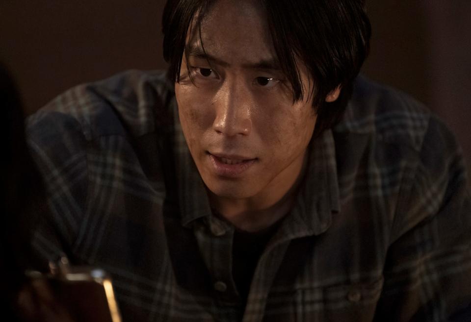 An Asian man with medium-length black hair is wearing a blue and brown plaid shirt and is holding a silver hipflask, his face is dirty.