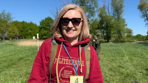 A blonde woman who appears in her forties smiles at the camera wearing sunglasses and a red hoodie, and camo trousers. 