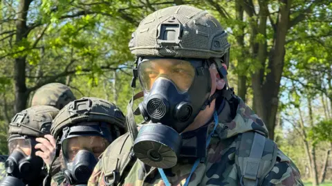 BBC A close-up of a young man wearing a gas mask, helmet and army camos. Behind him are three women wearing the same. The group is outdoors in a sunny field with trees. 