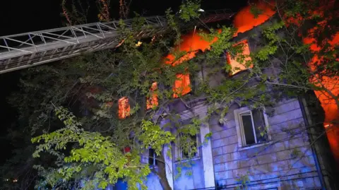 DSNS A residential building burns as a ladder extends from a fire truck that's out of view, with trees and foliage in the foreground, in Odesa on 1 May.