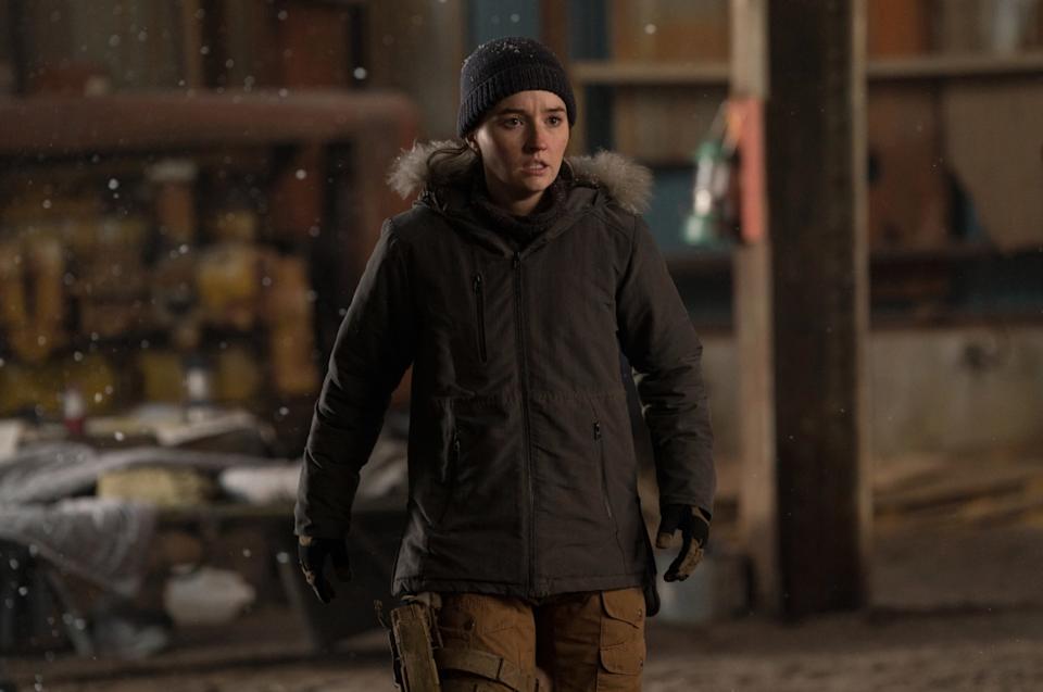 A young woman dressed in a black parka and wearing brown tactical trousers with a gun strapped to her left leg. She's wearing a black wooly hat. She's stood inside a warehouse.
