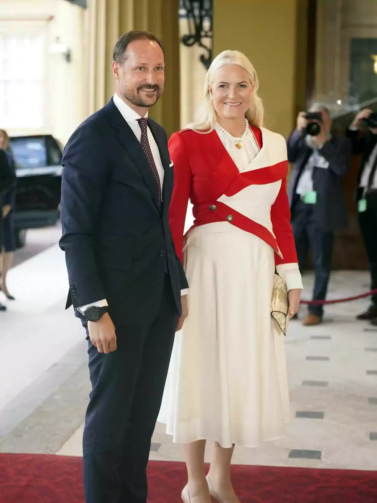 Norway's Haakon and elegantly dressed Mette-Marit pose for a photo at a formal event, with Haakon in a dark suit and Mette-Marit in a white dress and red jacket, while photographers capture the moment in the background.