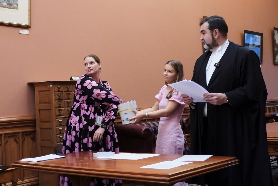 A woman wearing a floral dress draws from a cookie tin with a man wearing a black robe and a young girl holding the tin. 