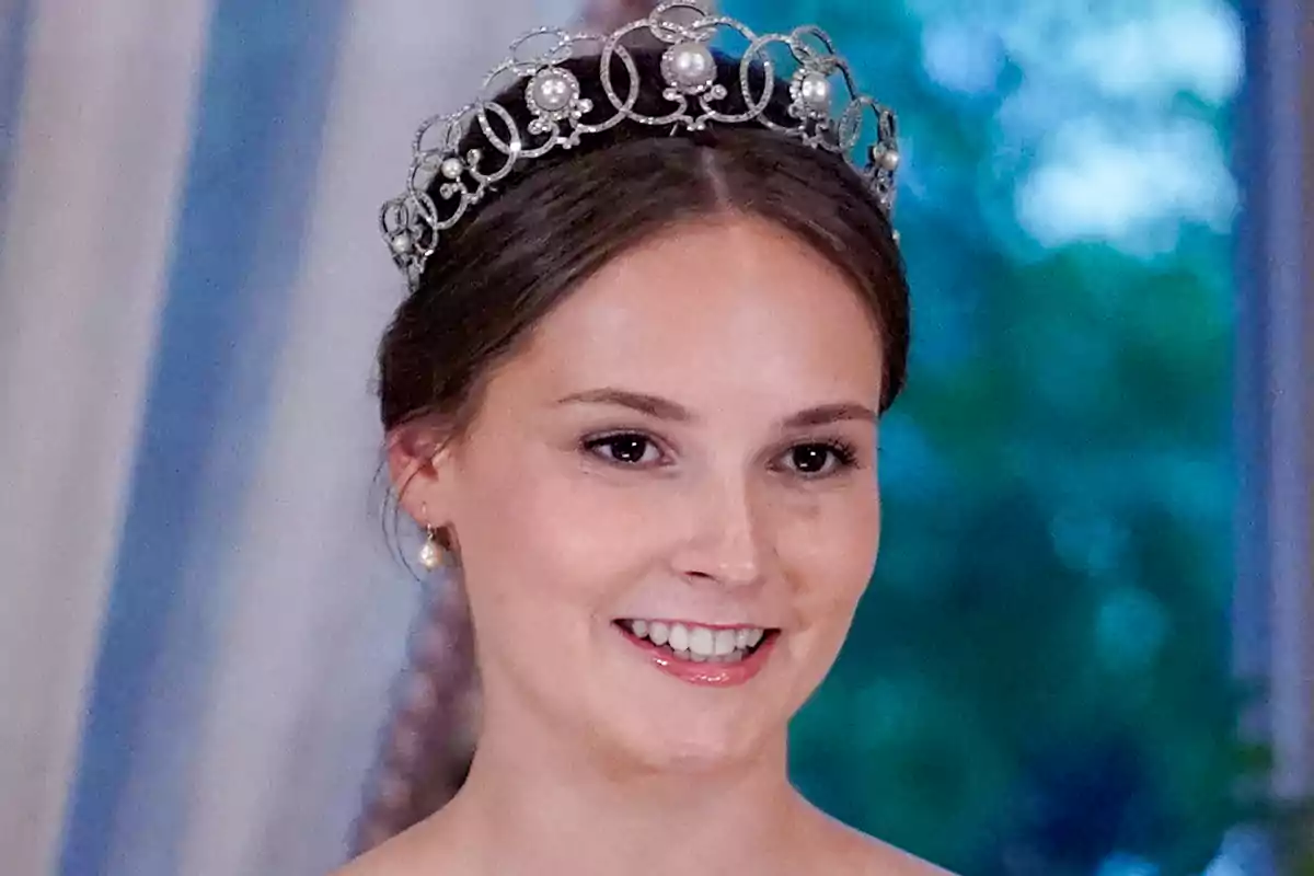 Princess Ingrid Alexandra, smiling, wears an elegant tiara with pearls and intricate details, set against a blurred background of blue and green tones.