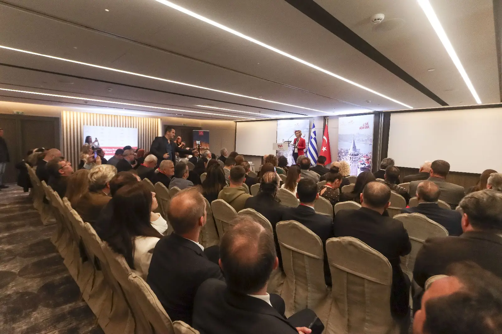 1.4 Million Turks Expected To Visit Greece In 2025 3 Attendees listening to a speaker at the Türkiye-Greece tourism workshop in a conference hall with Turkish and Greek flags.